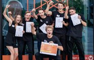Eine Gruppe junger Menschen posiert für die Kamera und hält Zertifikate in der einen Hand, während sie den anderen Arm in die Luft halten und jubeln