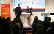 Zwei Menschen stehen auf der Bühne, das Publikum sitzt mit dem Rücken zur Kamera und hört zu