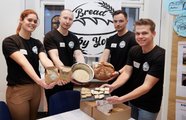 Vier Studierende stehen an ihrem Stand und halten ihr Produkt in die Kamera