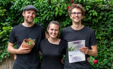 Die drei Gründer*innen von PRIMOZA stehen lächelnd nebeneinander und halten einen der wachsenden Kalender und einen Pflanzentopf mit einer Grünpflanze in der Hand.